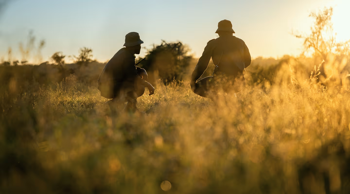 Two people in hats crouching in a field at sunset with tall grass and trees in the background.