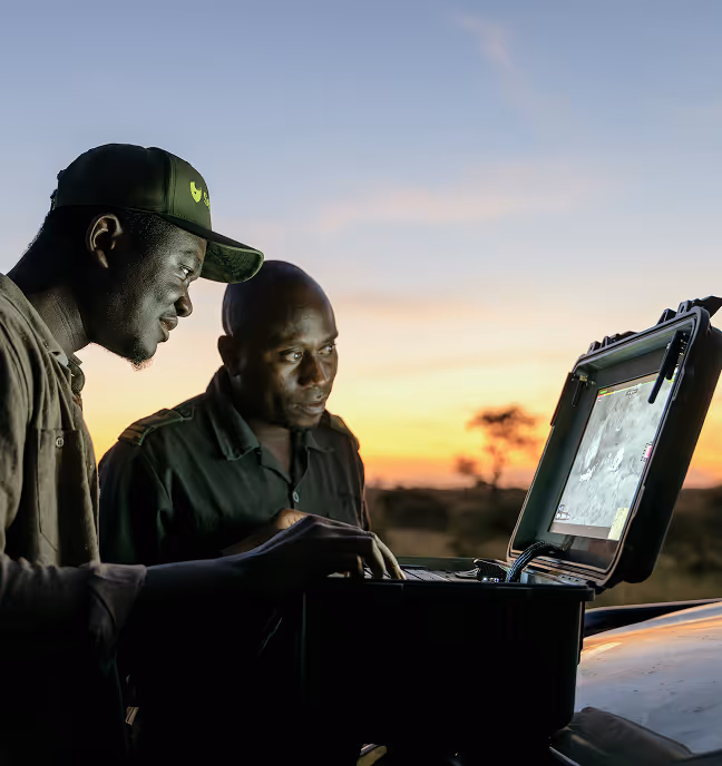 Two men outdoors at sunset examining data on a laptop mounted on a case.