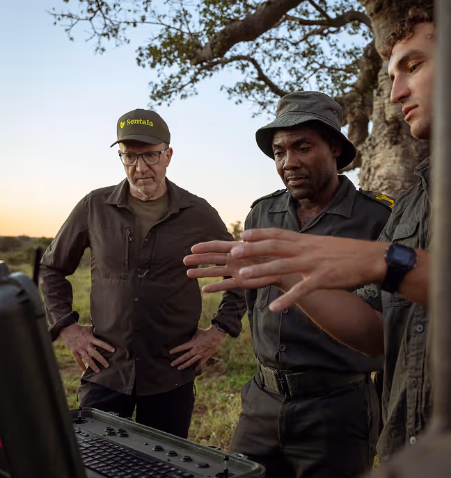 Three men outdoors in uniforms studying a screen by a tree at sunset during fieldwork.