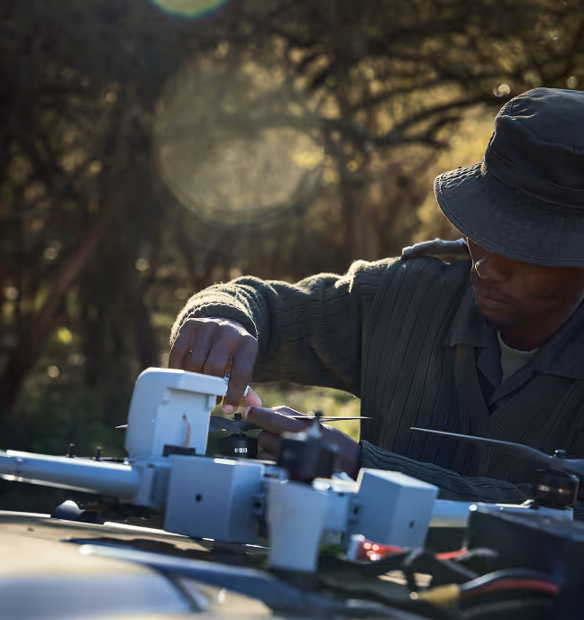 Man in a hat and green sweater inspecting or repairing a drone outdoors in a wooded area.