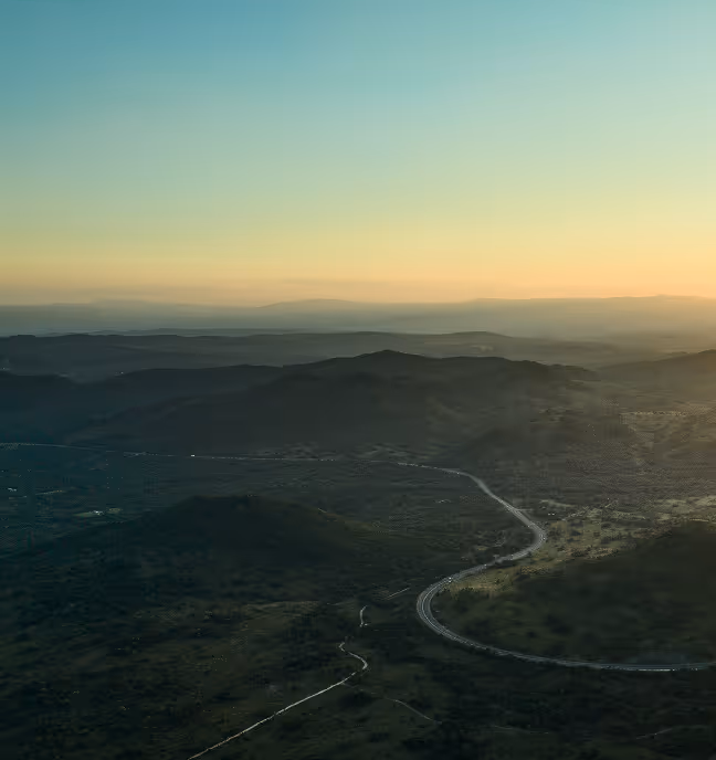 Curvy road winding through dark green hills under a clear sky at sunset.