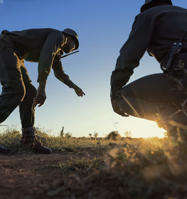 Two uniformed individuals with walkie-talkies crouching and pointing at the ground outdoors at sunset.