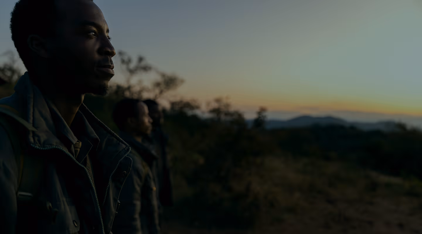 Three people standing outdoors at sunset, facing left with blurred trees and hills in the background.