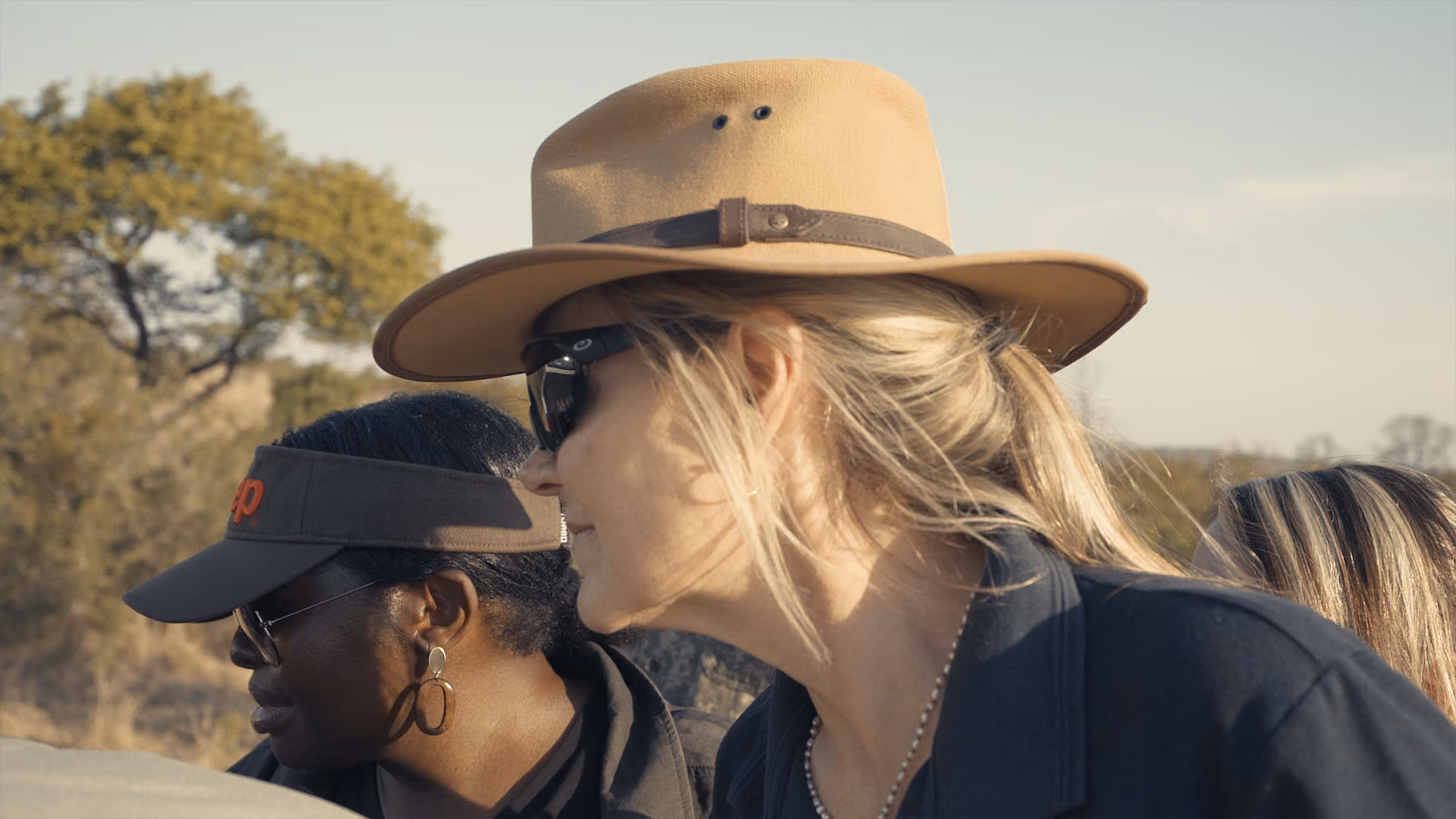 Two women outdoors, one wearing a tan hat and sunglasses, the other in a black cap and glasses.