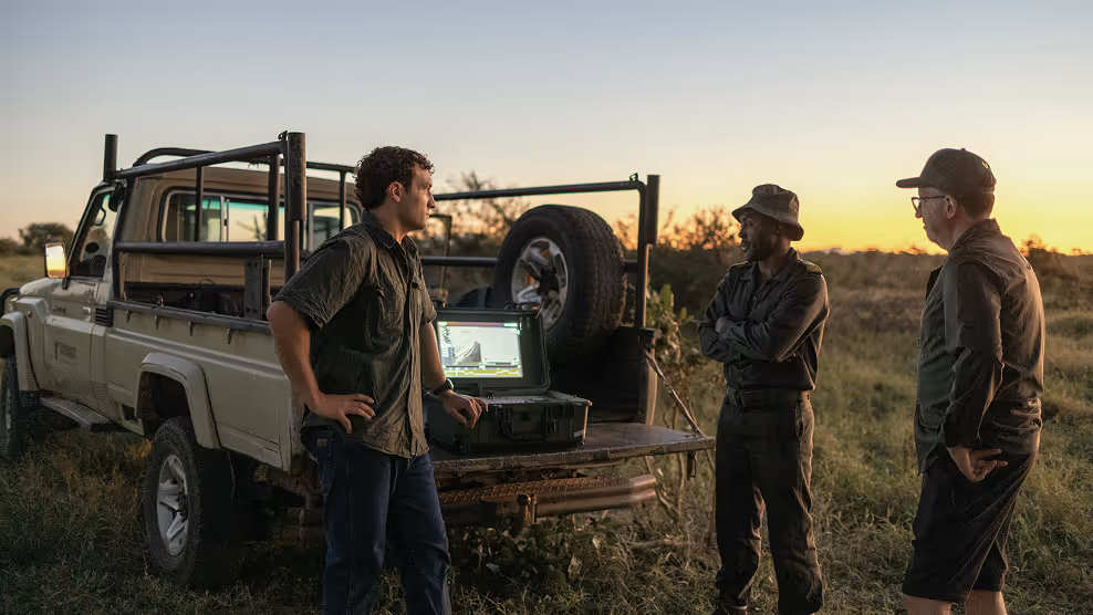 Three men standing by a pickup truck with open tailgate watching a screen at sunset outdoors.