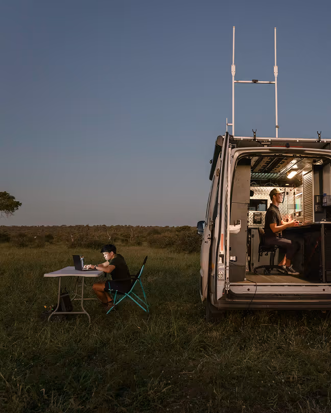 Two people working at laptops in a grass field, one inside a van and one at a folding table nearby at dusk.