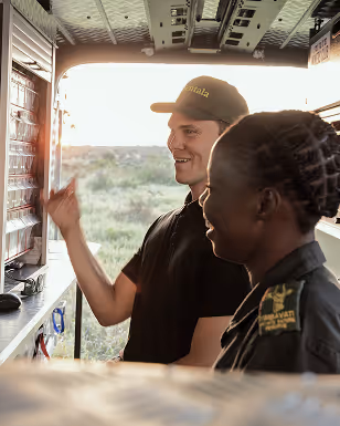Two people smiling inside a vehicle, one pointing at a control panel on the left side.