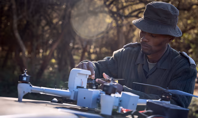 Man wearing hat adjusting drone outdoors with trees blurred in background at sunset.